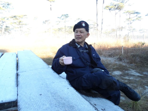 พล.ต.ต.ชฎิล พรหมไพบูลย์ ผู้บังคับการตำรวจภูธรจังหวัดพิษณุโลก นั่งดื่มกาแฟ บนโต๊ะกลางแจ้งที่มีแม่คะนิ้งเกาะพราวเต็มโต๊ะ บน “ภูสอยดาว”