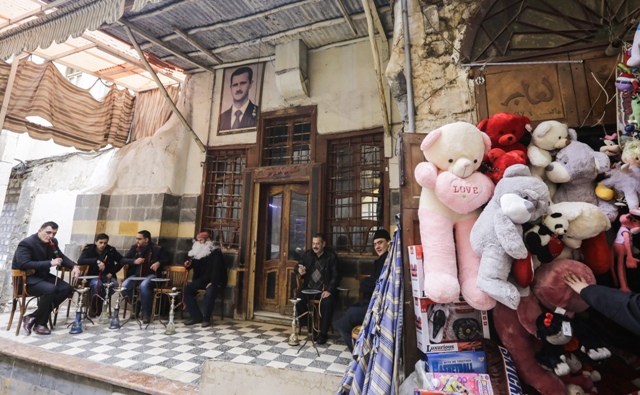 ชาวซีเรียนั่งอยู่ด้านนอกคาเฟ่แห่งหนึ่งที่ติดรูปประธานาธิบดี บาชาร์ อัล-อัสซาด ในขณะที่ร้านค้าข้างๆ ขายของขวัญวาเลนไทน์ในพื้นที่เก่าแก่ของกรุงดามัสกัส (12 ก.พ.)