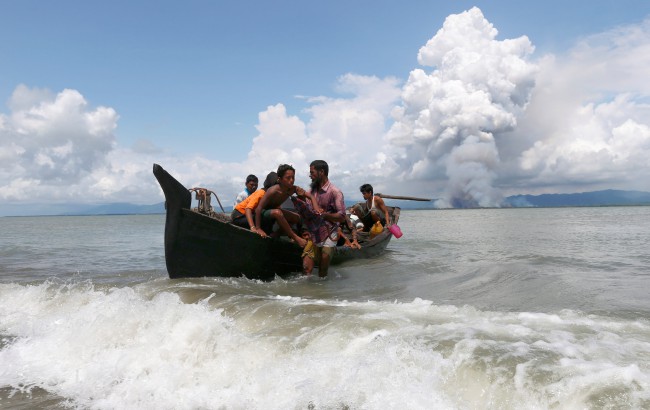กลุ่มควันขนาดใหญ่พวยพุ่งขึ้นอยู่ด้านหลังในฝั่งพม่า ขณะเรือที่บรรทุกผู้ลี้ภัยโรฮิงญาข้ามอ่าวเบงกอลมาถึงฝั่งบังกลาเทศ แต่ขณะเดียวกันก็มีเรือของผู้ลี้ภัยหลายลำที่ไปไม่ถึงฝั่งเกิดพลิกคว่ำอับปางกลางทาง. -- Reuters/Denis Balibouse.