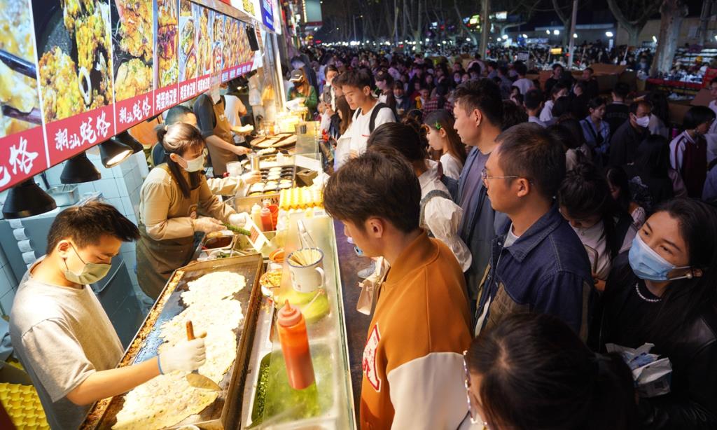 ลูกค้าหนุ่มสาวรอซื้ออาหารแผงลอยที่ตลาดกลางคืนในเมืองเจิ้งโจว มณฑลเหอหนันทางภาคกลางของจีนเมื่อวันที่ 2 เม.ย.2566 โดยหลายมณฑลและเมืองต่างๆ ทั่วประเทศเร่งส่งเสริมการบริโภคของประชาชนในช่วงที่สภาพอากาศเริ่มอบอุ่นในขณะนี้ - ภาพ : VCG